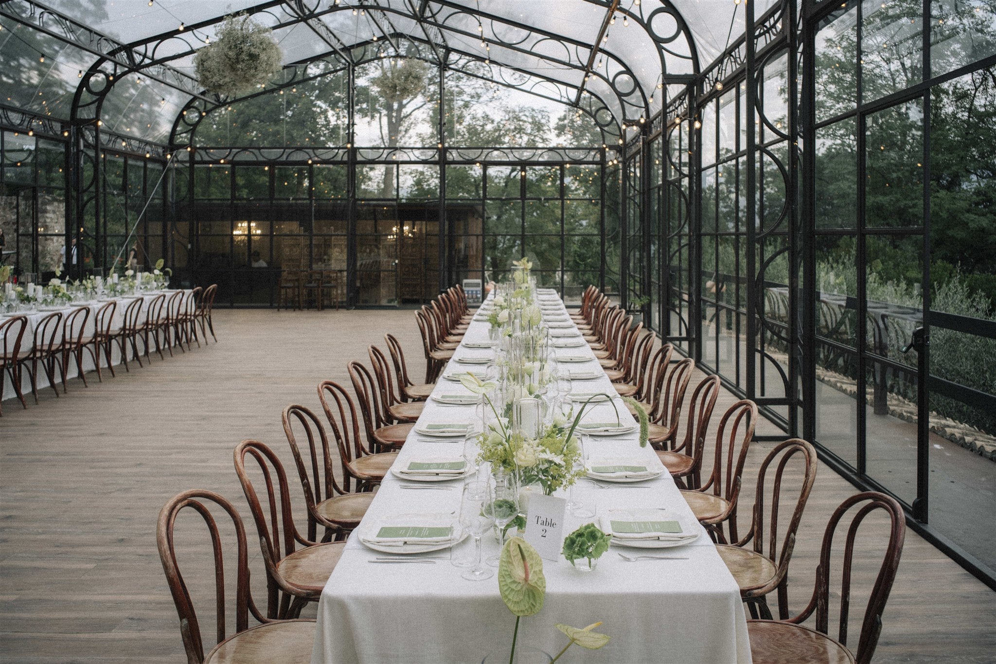 Festliche Tafel mit rustikalen Holzstühlen und eleganter, grüner Tischdekoration in einem Glaspavillon.