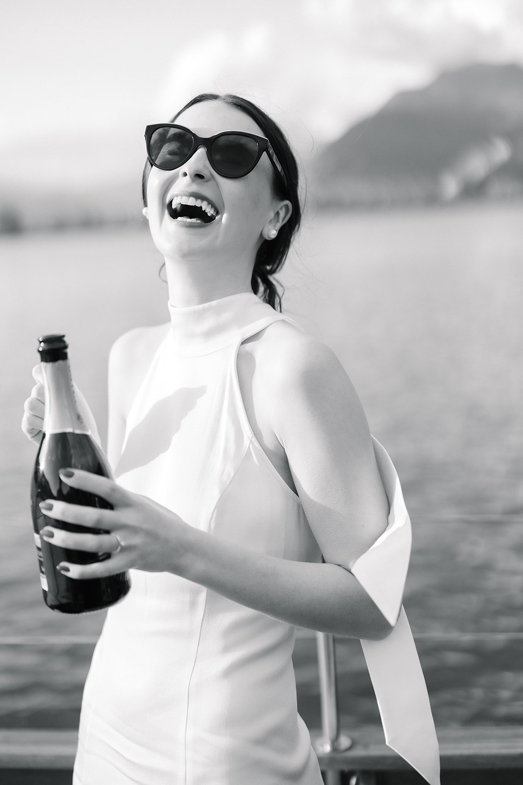 Braut mit Sonnenbrille und Champagnerflasche, strahlend vor Freude, mit einer malerischen Landschaft im Hintergrund