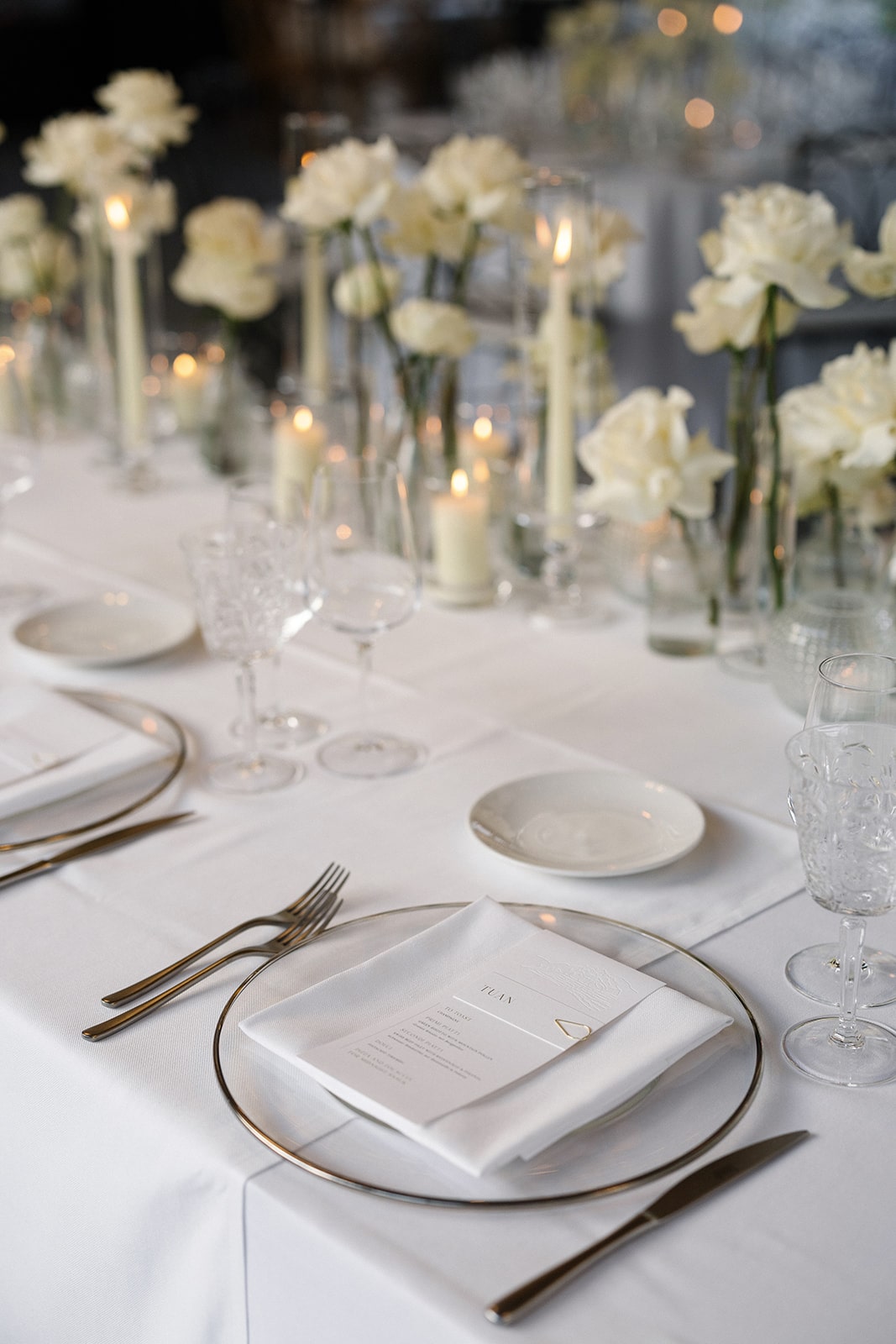 Großes Elegante Hochzeitstafel mit weißen Rosen, Kerzen und gläsernen Details, die für ein romantisches Ambiente sorgen.