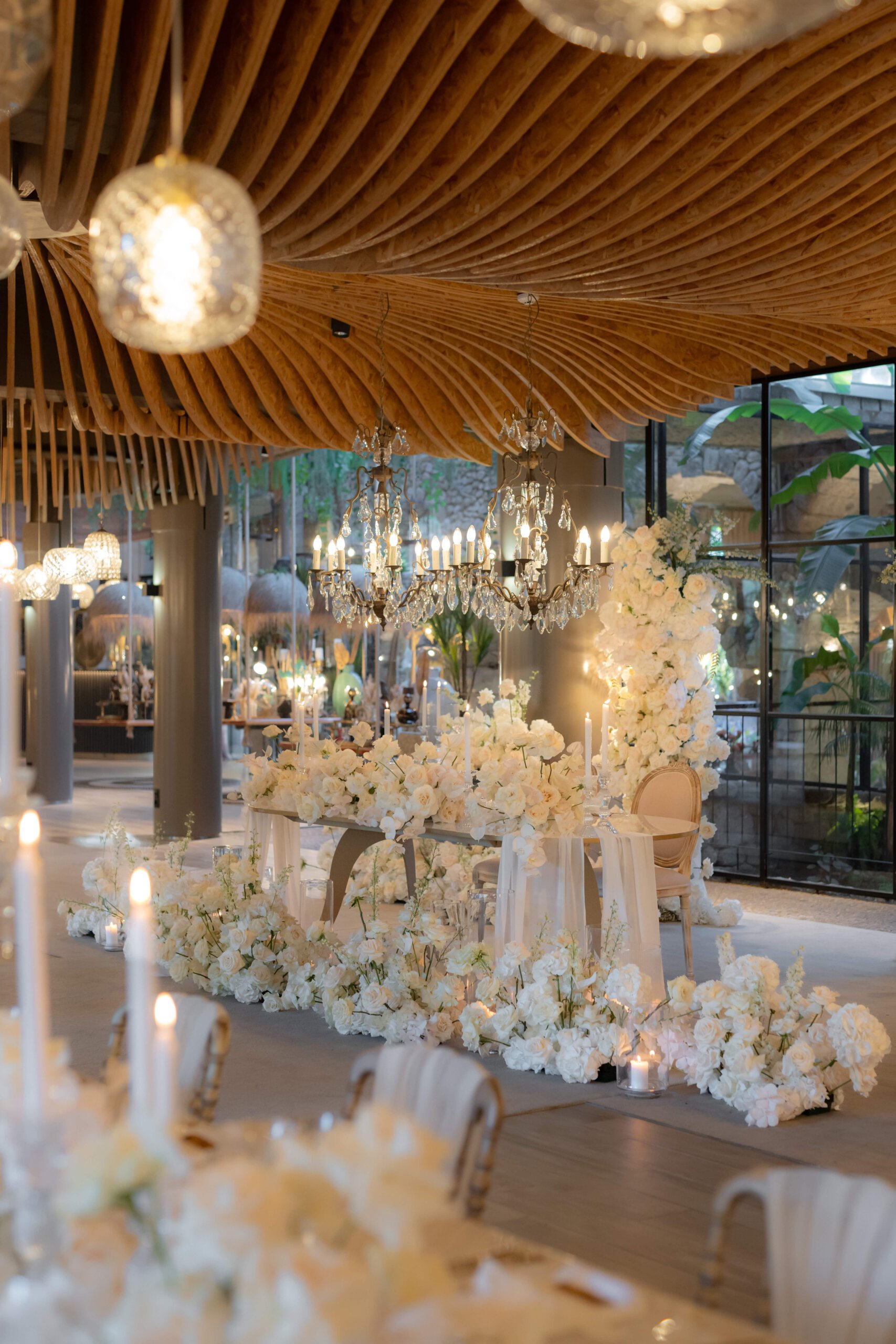 Elegant dekorierter Hochzeitsempfang mit weißen Blumenarrangements und funkelnden Kronleuchtern unter einer modernen, wellenförmigen Holzdecke, die eine luxuriöse Atmosphäre schafft.