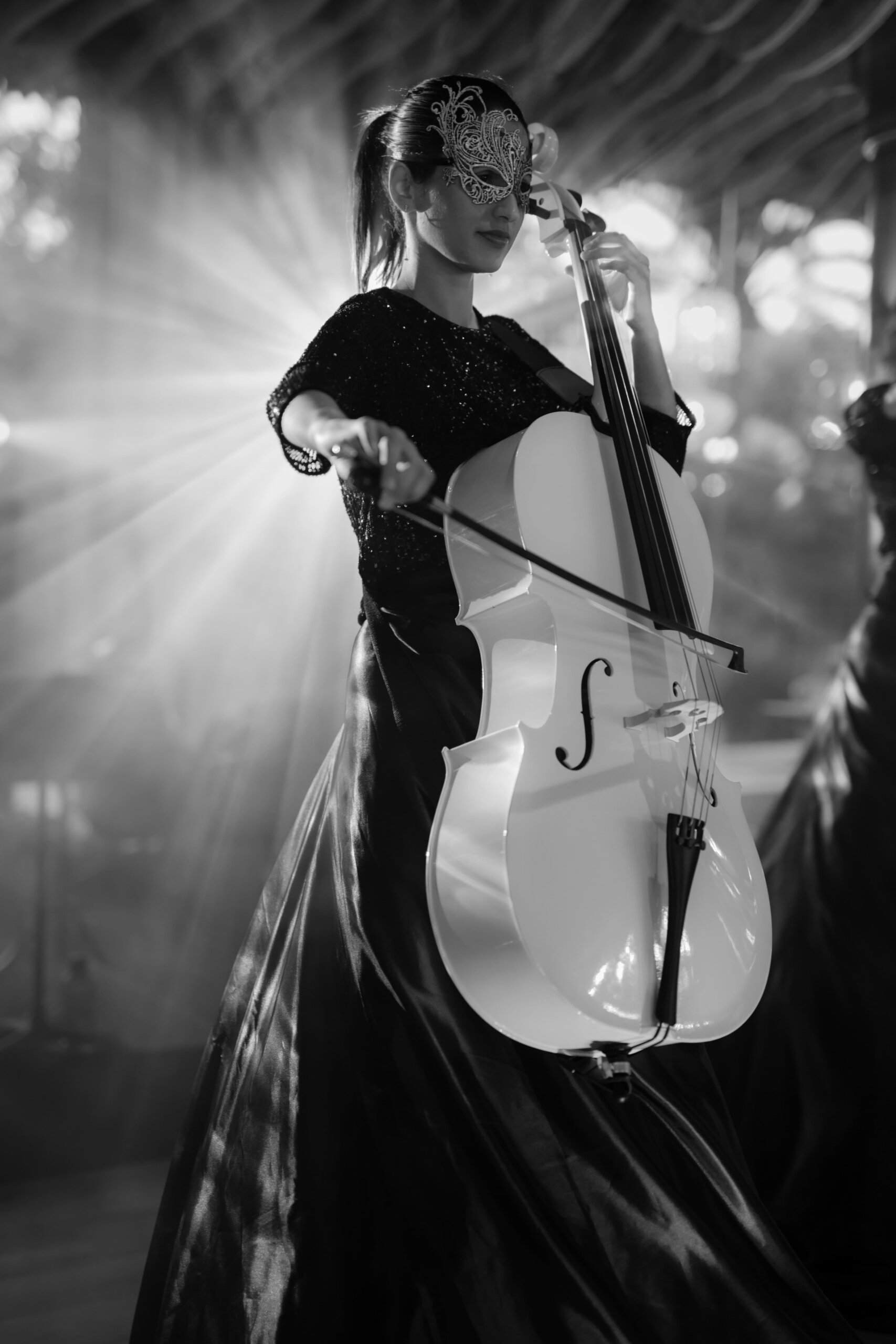 Schwarz-Weiß-Aufnahme einer Musikerin, die ein weißes Cello spielt, mit einer kunstvollen Augenmaske und dramatischer Bühnenbeleuchtung, die eine magische Atmosphäre erzeugt.