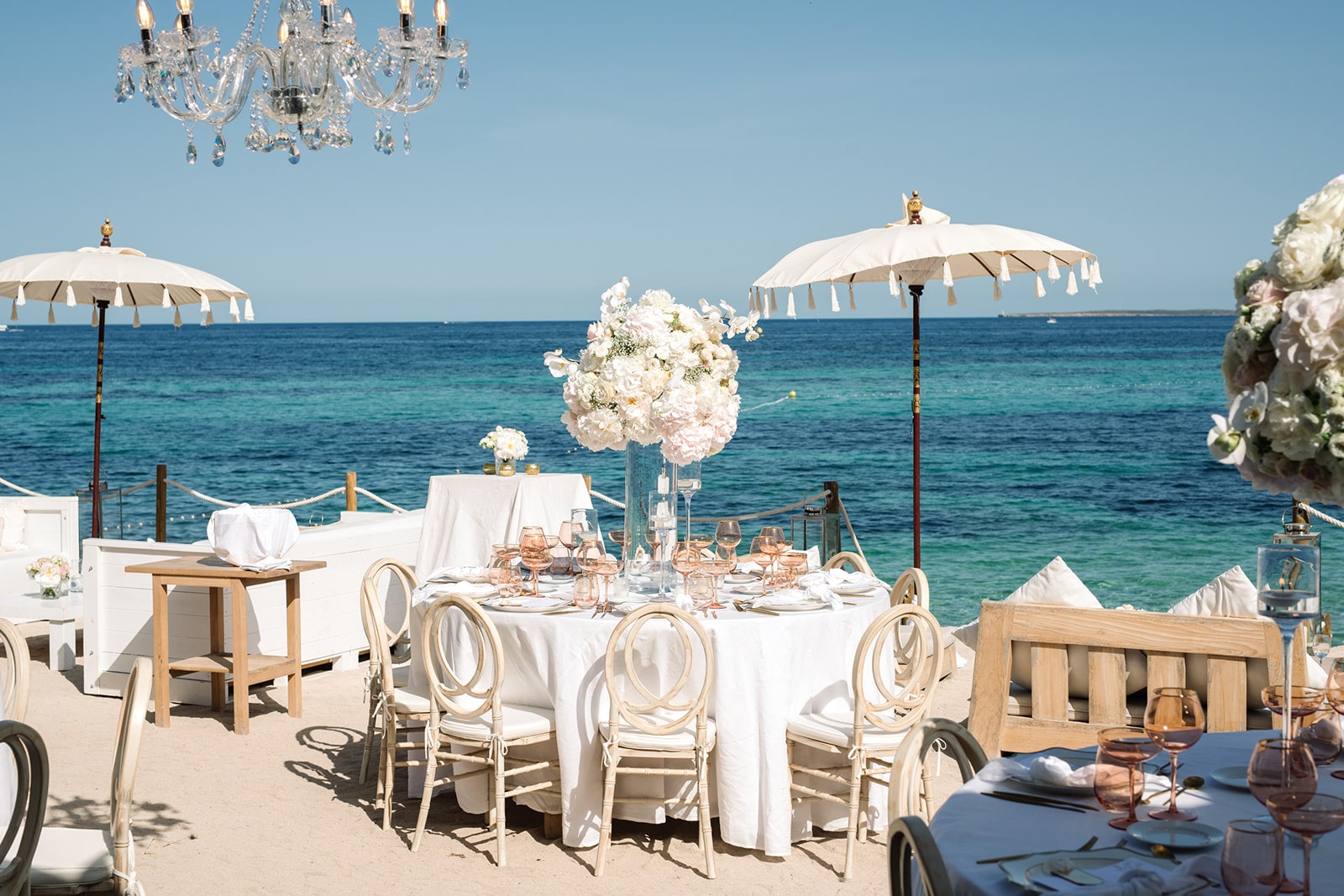 Luxuriöse Strandhochzeit mit Kronleuchter, eleganten Tischen und einem klaren blauen Himmel.