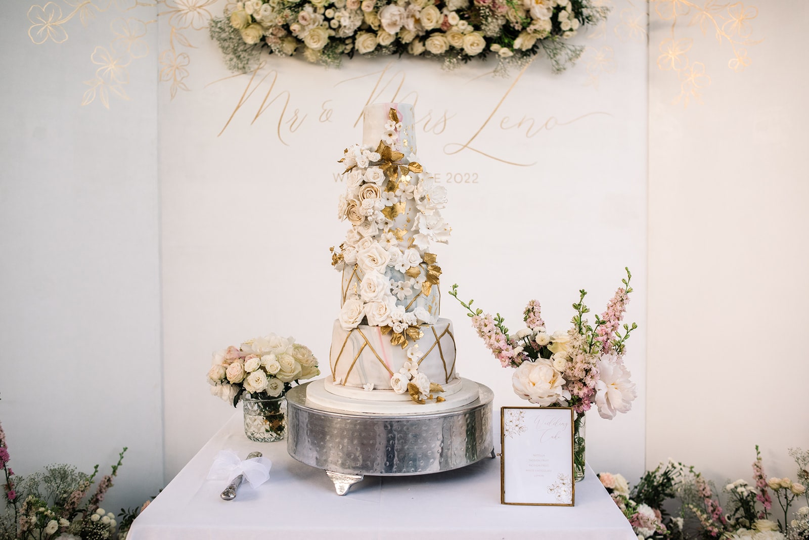 Prächtige Hochzeitstorte in Weiß und Gold mit kunstvoller Blumendekoration, umgeben von floralen Arrangements.