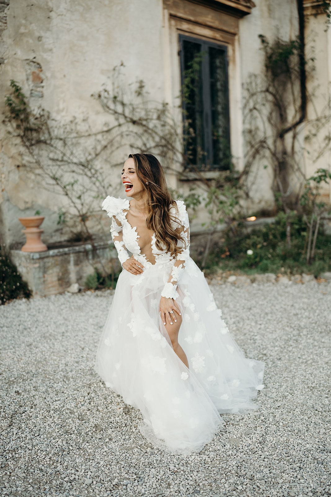 Braut strahlt und lacht in einem traumhaften Hochzeitskleid mit floralen Details vor einer alten Villa.