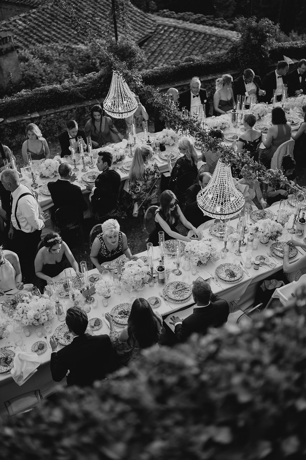 Luftaufnahme einer eleganten Hochzeitstafel im Freien, umgeben von stilvoll gekleideten Gästen, funkelnden Kronleuchtern und prachtvollen Blumenarrangements.