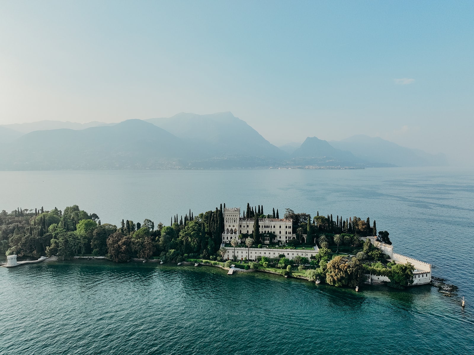 Luftaufnahme einer beeindruckenden italienischen Inselvilla, umgeben von tiefblauem Wasser und malerischen Bergen im Hintergrund.