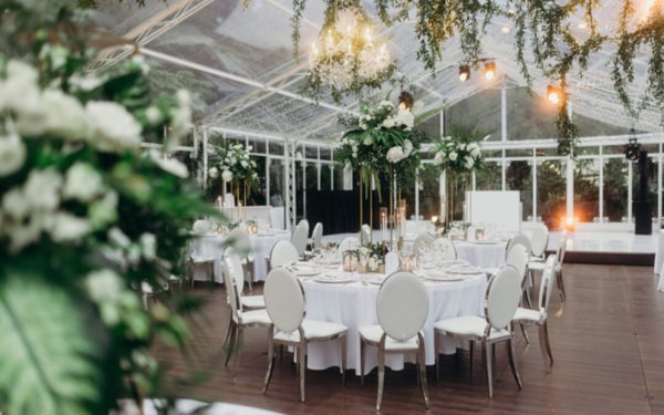 Blick auf ein prachtvolles Hochzeitssetup in einem gläsernen Festzelt, dekoriert mit weißen Blumen, hohen Vasen und hängendem Efeu für eine luxuriöse Atmosphäre.