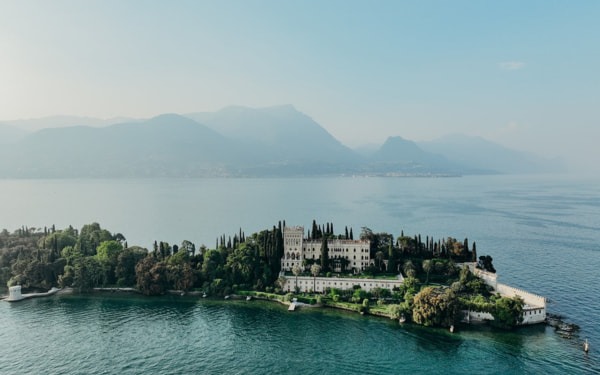 Luftaufnahme einer beeindruckenden italienischen Inselvilla, umgeben von tiefblauem Wasser und malerischen Bergen im Hintergrund.