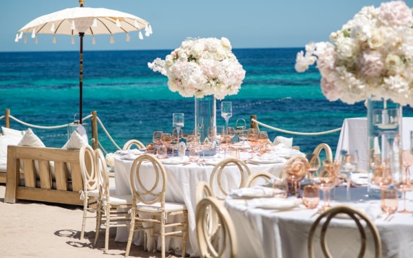 Elegante Hochzeitstafel am Strand in Ibiza mit luxuriösen Blumengestecken und Kerzen, die eine romantische Atmosphäre schaffen