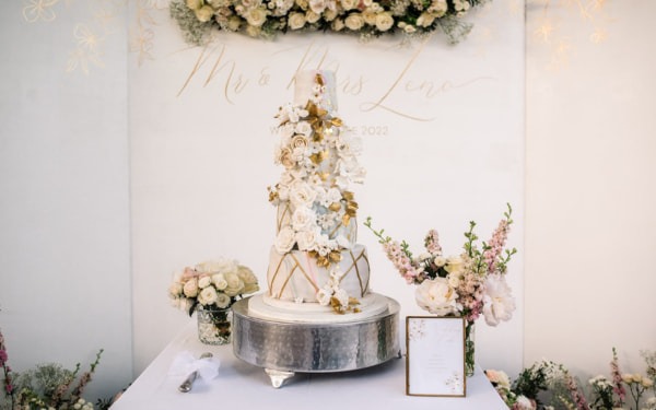 Prächtige Hochzeitstorte in Weiß und Gold mit kunstvoller Blumendekoration, umgeben von floralen Arrangements.