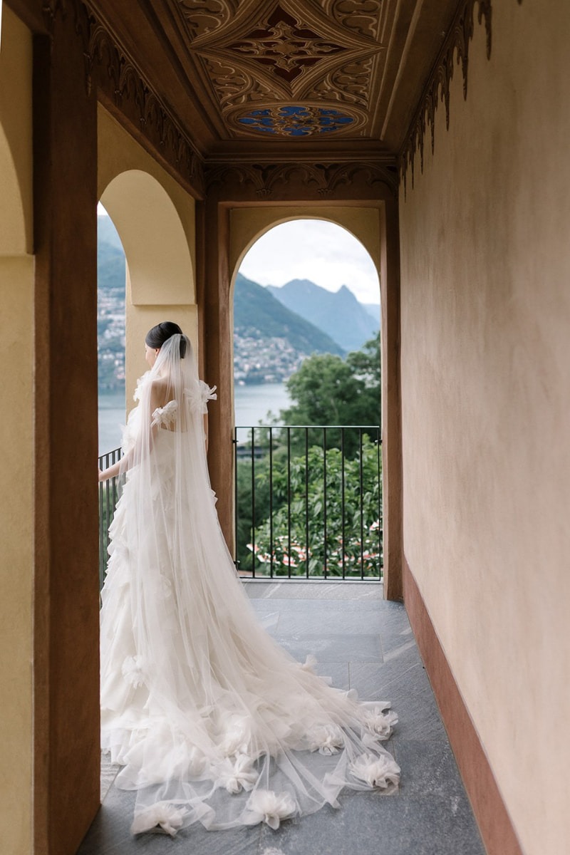 Braut in einem eleganten Hochzeitskleid, posierend auf einem Balkon mit Blick auf einen idyllischen See und Berge