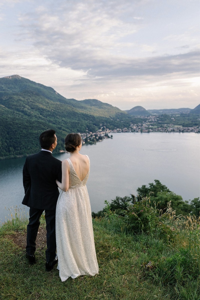 Braut und Bräutigam stehen auf einer Wiese mit Blick auf einen malerischen See und die umliegenden Hügel in der Abenddämmerung.